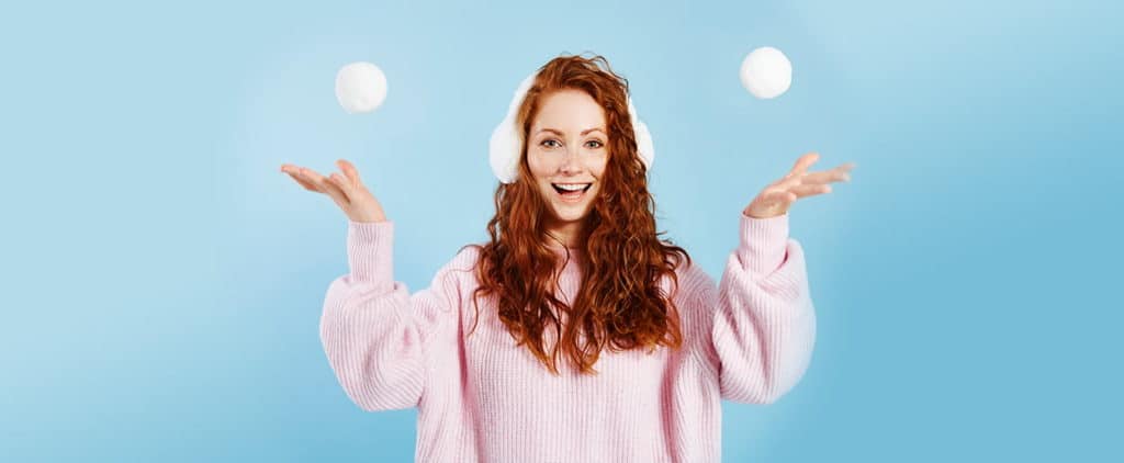 Woman with red hair wearing earmuffs and pink sweater tossing snowballs up in the air